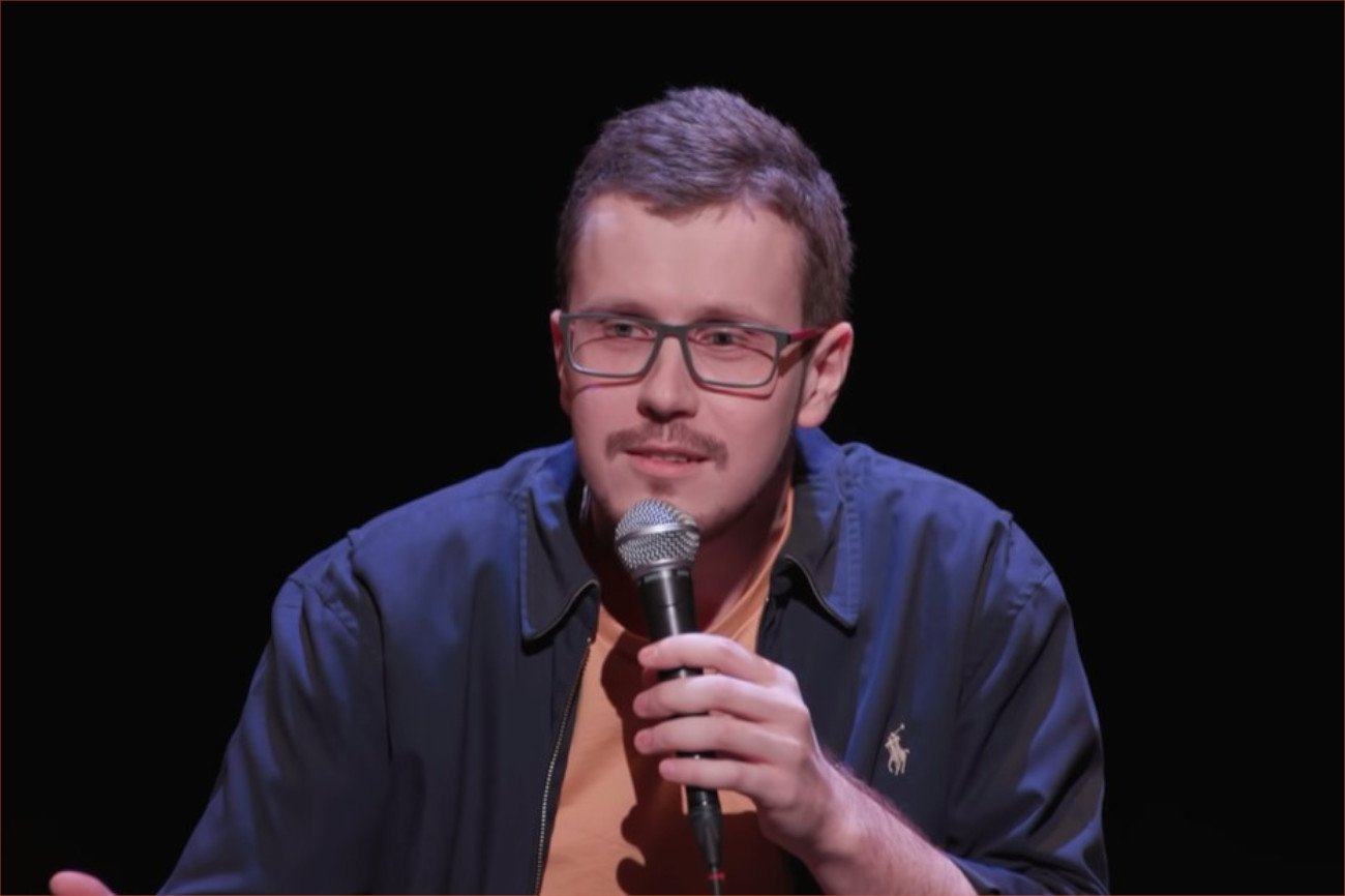 Na zdjęciu widnieje mężczyzna w okularach, który trzyma mikrofon i mówi na scenie. W tle ciemne tło.